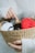 A woven basket held in two hands contains several balls of yarn in various colors, including red, gray, white, and black. On top of the yarn, a small figurine of a yellow and white animal with small ears and eyes sits, partially in a small cardboard box. The index finger of the right hand is pointing towards this figurine. The person holding the basket is wearing a white ribbed sweater.