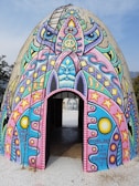 A dome-shaped structure covered in vibrant, psychedelic art. The artwork includes various symbols such as peace signs, stars, and abstract faces in vivid colors like pink, blue, and yellow. There is a ladder leading to the top, and an opening at the base provides a view inside.
