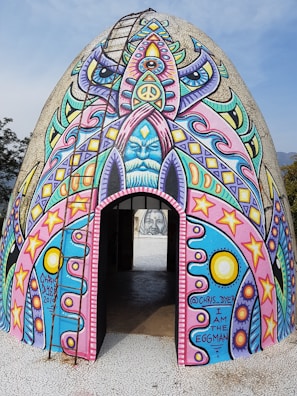 A dome-shaped structure covered in vibrant, psychedelic art. The artwork includes various symbols such as peace signs, stars, and abstract faces in vivid colors like pink, blue, and yellow. There is a ladder leading to the top, and an opening at the base provides a view inside.