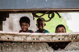 Three children are peering through the gap of a large piece of machinery or construction equipment. They have cheerful expressions, suggesting a sense of playfulness and curiosity.