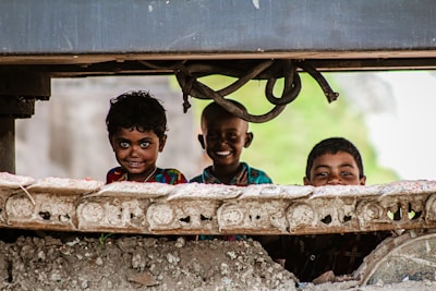 Three children are peering through the gap of a large piece of machinery or construction equipment. They have cheerful expressions, suggesting a sense of playfulness and curiosity.