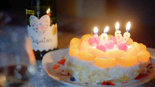 A birthday cake topped with slices of fruit and four lit candles shaped as the numbers one, zero, six, and eight. The background includes a bottle labeled 'Piquitos' with a colorful heart design. The cake is on a plate with a decorative pattern, and the setting appears to be softly lit, creating a warm ambiance.