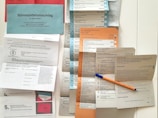 A collection of voting ballots and official documents are neatly arranged on a flat surface. The documents include multiple forms and ballots, some with checkboxes and political party information in German. A blue pen with an orange body is placed on top, likely used for filling out the forms.
