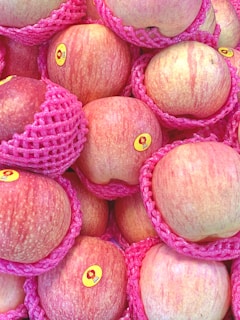 A pile of fresh apples encased in pink foam netting, each with a small sticker on them. The apples have a red and yellow streaked appearance and the foam offers protection. The scene emphasizes the organized presentation of the fruit.