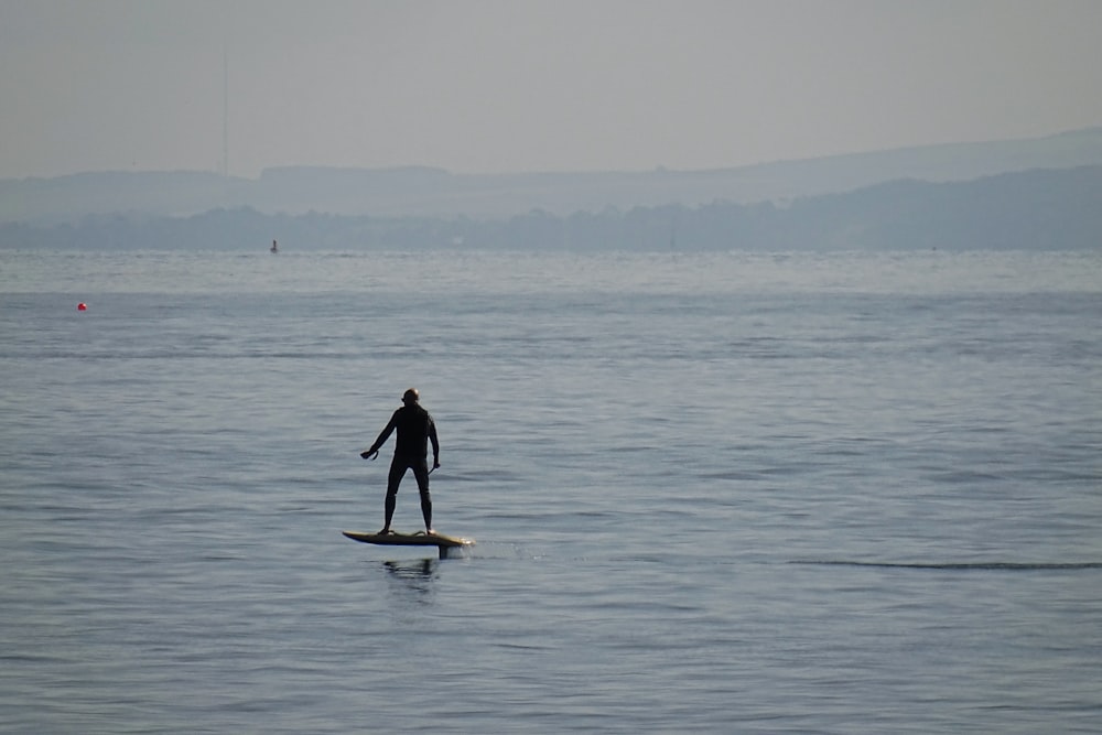 낮 동안 바다에서 하얀 서핑 보드에 서있는 검은 잠수복을 입은 남자