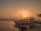 A scenic view of a marina at sunset with several boats docked near the shoreline. The sky is a warm orange hue, casting a gentle glow on the water. Palm trees can be seen in the background, adding a tropical feel to the scene. A small sign by the dock provides information about boat tours.