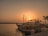 A scenic view of a marina at sunset with several boats docked near the shoreline. The sky is a warm orange hue, casting a gentle glow on the water. Palm trees can be seen in the background, adding a tropical feel to the scene. A small sign by the dock provides information about boat tours.