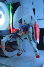 A model of a Gundam robot stands in front of a gaming computer setup. The robot is detailed, featuring red, blue, yellow, and white coloring, and is equipped with a shield and weapon. The background consists of computer hardware components illuminated by colorful LED lights, adding an ambient glow to the scene.