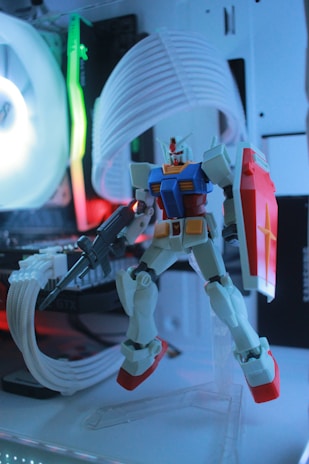 A model of a Gundam robot stands in front of a gaming computer setup. The robot is detailed, featuring red, blue, yellow, and white coloring, and is equipped with a shield and weapon. The background consists of computer hardware components illuminated by colorful LED lights, adding an ambient glow to the scene.