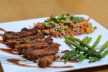 A plate of food featuring sliced grilled meat with a glaze, a mixed vegetable and grain salad, and several stalks of asparagus. The dish is arranged neatly on a white plate with decorative sauce drizzle.