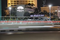 A nighttime cityscape featuring a hotel with an illuminated sign, surrounded by tropical plants and palm trees. A circular fountain with water jets creates arcs in front of the building. Light trails from vehicles indicate motion on the road.