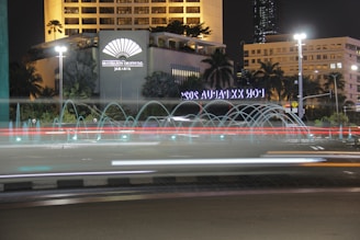 A nighttime cityscape featuring a hotel with an illuminated sign, surrounded by tropical plants and palm trees. A circular fountain with water jets creates arcs in front of the building. Light trails from vehicles indicate motion on the road.