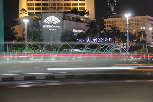 A nighttime cityscape featuring a hotel with an illuminated sign, surrounded by tropical plants and palm trees. A circular fountain with water jets creates arcs in front of the building. Light trails from vehicles indicate motion on the road.