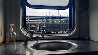 A train carriage washbasin with a metal sink, a chrome faucet, and a small bottle of liquid soap on the left. Through the window, a railway station platform is visible with a freight train stopped outside. The scene has an industrial and utilitarian feel.
