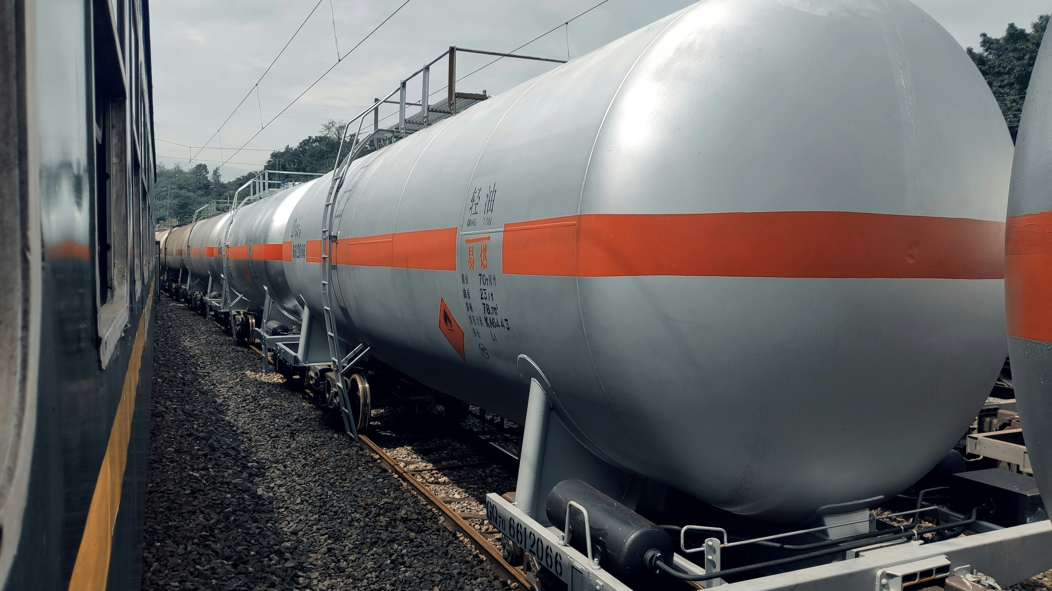 A series of industrial tank cars are lined up on railway tracks. These cylindrical tanks, painted in silver with an orange stripe, appear to be used for transporting liquid chemicals or fuels. The scene includes a train on the adjacent track, and the background shows some trees and electrical wires.