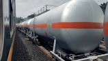 A series of industrial tank cars are lined up on railway tracks. These cylindrical tanks, painted in silver with an orange stripe, appear to be used for transporting liquid chemicals or fuels. The scene includes a train on the adjacent track, and the background shows some trees and electrical wires.