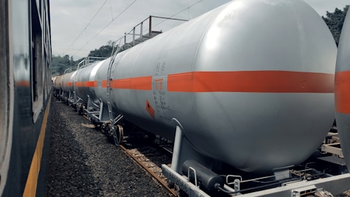 A series of industrial tank cars are lined up on railway tracks. These cylindrical tanks, painted in silver with an orange stripe, appear to be used for transporting liquid chemicals or fuels. The scene includes a train on the adjacent track, and the background shows some trees and electrical wires.