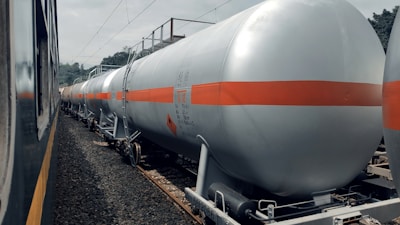A series of industrial tank cars are lined up on railway tracks. These cylindrical tanks, painted in silver with an orange stripe, appear to be used for transporting liquid chemicals or fuels. The scene includes a train on the adjacent track, and the background shows some trees and electrical wires.