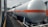 A series of industrial tank cars are lined up on railway tracks. These cylindrical tanks, painted in silver with an orange stripe, appear to be used for transporting liquid chemicals or fuels. The scene includes a train on the adjacent track, and the background shows some trees and electrical wires.