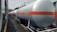 A series of industrial tank cars are lined up on railway tracks. These cylindrical tanks, painted in silver with an orange stripe, appear to be used for transporting liquid chemicals or fuels. The scene includes a train on the adjacent track, and the background shows some trees and electrical wires.