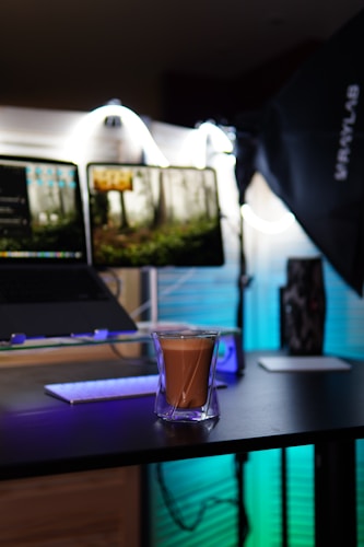 A modern workspace setup with dual monitors displaying a forest scene. A glass of coffee is set on a dark surface in the foreground. LED light strips create an ambient glow with blue and white tones. A camera light with a softbox is positioned to the side, and a sleek keyboard is visible. Cables are neatly managed, enhancing a tech-savvy atmosphere.