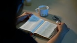 A person is reading an open book, possibly a Bible, placed on a table. Beside the book, there is a white mug with a floral pattern and a pair of glasses. The lighting is dim, creating a calm and peaceful atmosphere.