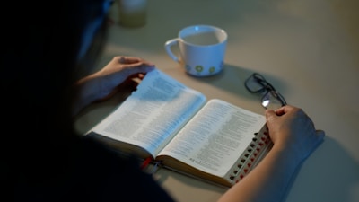 A person is reading an open book, possibly a Bible, placed on a table. Beside the book, there is a white mug with a floral pattern and a pair of glasses. The lighting is dim, creating a calm and peaceful atmosphere.