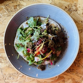 A plate of salad consisting of leafy greens, possibly arugula, mixed with other colorful ingredients such as diced tomatoes and some crumbled, herb-coated bits. The salad is served on a flatbread and garnished with dressing and spices. The plate is set on a textured wooden surface.