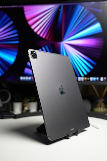 A sleek tablet with an Apple logo, positioned on a stand, with a vibrant abstract design on a large screen in the background. The setting appears to be a modern desk with minimalistic decor.