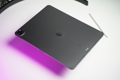 A sleek, dark-colored tablet lies on a surface with a distinctive groove design on the back, accompanied by a stylus nearby. Soft purple lighting casts a gradient effect across the surface.