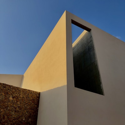 A modern architectural structure featuring sharp geometric lines and a mix of materials such as concrete and stone. The building has a tall, rectangular form with a prominent open section near the top that contrasts against a clear blue sky.