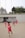 Several children in red sports uniforms are actively participating in a cone drill on a large concrete surface. The background features a modern white building, trees, and a few people sitting or standing near the structure.