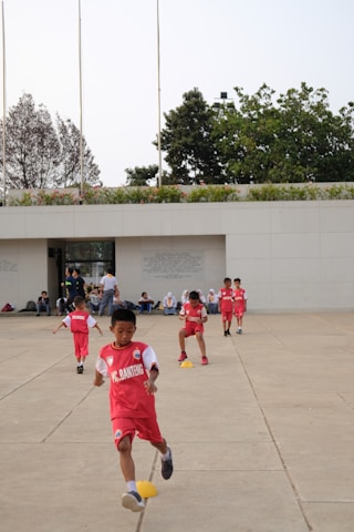 Several children in red sports uniforms are actively participating in a cone drill on a large concrete surface. The background features a modern white building, trees, and a few people sitting or standing near the structure.