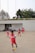 Several children in red sports uniforms are actively participating in a cone drill on a large concrete surface. The background features a modern white building, trees, and a few people sitting or standing near the structure.