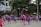 A group of basketball players in purple and red uniforms are actively engaged in a game on an outdoor court surrounded by spectators. The players are captured mid-action, with one player jumping and reaching for the ball. The background includes trees, a fence, and people watching from the sidelines.