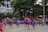 A group of basketball players in purple and red uniforms are actively engaged in a game on an outdoor court surrounded by spectators. The players are captured mid-action, with one player jumping and reaching for the ball. The background includes trees, a fence, and people watching from the sidelines.