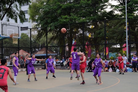 A group of basketball players in purple and red uniforms are actively engaged in a game on an outdoor court surrounded by spectators. The players are captured mid-action, with one player jumping and reaching for the ball. The background includes trees, a fence, and people watching from the sidelines.