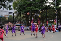A group of basketball players in purple and red uniforms are actively engaged in a game on an outdoor court surrounded by spectators. The players are captured mid-action, with one player jumping and reaching for the ball. The background includes trees, a fence, and people watching from the sidelines.