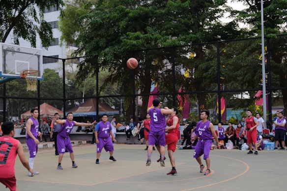 A group of basketball players in purple and red uniforms are actively engaged in a game on an outdoor court surrounded by spectators. The players are captured mid-action, with one player jumping and reaching for the ball. The background includes trees, a fence, and people watching from the sidelines.