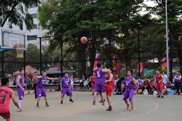 A group of basketball players in purple and red uniforms are actively engaged in a game on an outdoor court surrounded by spectators. The players are captured mid-action, with one player jumping and reaching for the ball. The background includes trees, a fence, and people watching from the sidelines.