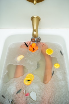 A close-up view of a luxurious bathtub filled with water and bubbles. Inside the tub, slices of oranges and leaves are floating, creating a spa-like atmosphere. The bronze faucet adds a vintage touch, and a person's feet are partially submerged.