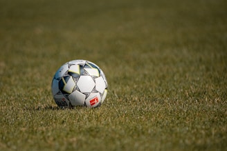 A soccer ball resting on a grassy field, featuring a modern design with geometric patterns and a small red logo.