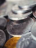 A close-up view of a pile of coins, primarily metallic in appearance with distinct engravings and details visible on the surface.