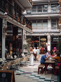 An indoor market or shopping arcade with ornate, decorative ironwork and high ceilings. People are dining and walking, enjoying the ambiance. There are tables and chairs set for dining along the passageway.