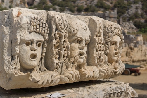 A stone relief features three ancient theatrical masks, intricately carved with detailed facial expressions. The masks are positioned side by side on a weathered stone block. In the background, there is a blurred landscape of stone ruins, indicating an archaeological site.