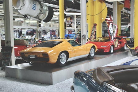 A collection of vintage cars on display in an automotive showroom, featuring a prominent yellow sports car and a red sports car with its gull-wing doors open. The cars are showcased on elevated platforms, surrounded by other classic car models and various automobile memorabilia.