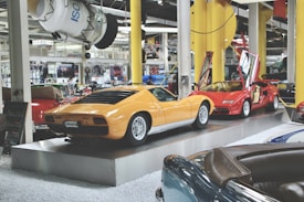 A collection of vintage cars on display in an automotive showroom, featuring a prominent yellow sports car and a red sports car with its gull-wing doors open. The cars are showcased on elevated platforms, surrounded by other classic car models and various automobile memorabilia.
