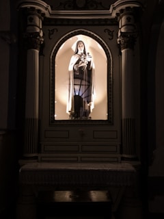 A religious statue is housed within an ornate alcove. The statue depicts a figure in robes, illuminated by soft lighting, creating a serene atmosphere. The alcove is framed with detailed architectural elements including columns and decorative moldings.