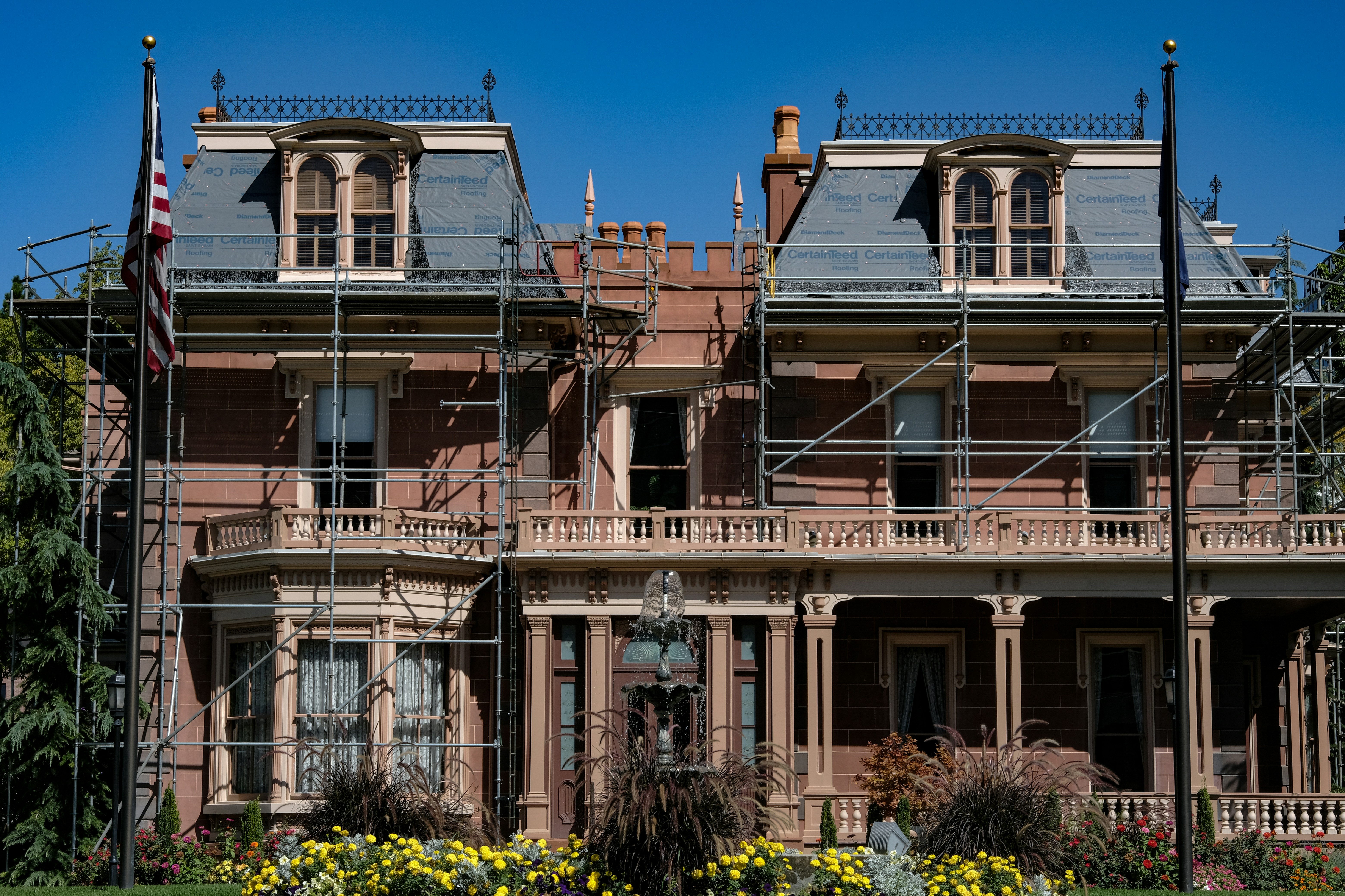 A large, historic-looking house with scaffolding around its structure, suggesting renovations or repairs. The building features ornate architectural details and is surrounded by vibrant greenery and colorful flowers. An American flag is visible on a flagpole in front of the house.
