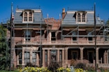 A large, historic-looking house with scaffolding around its structure, suggesting renovations or repairs. The building features ornate architectural details and is surrounded by vibrant greenery and colorful flowers. An American flag is visible on a flagpole in front of the house.
